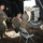 Military personnel in MARPAT camouflage uniforms are operating communications equipment inside a field command post tent. Two soldiers are seated at a table with radio and electronic equipment while a third stands observing. The setting appears to be an outdoor field exercise or training operation.