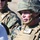 Two U.S. Marines in full combat gear are visible in the foreground and background. The primary subject wears a name tape reading 'RUSSO' with rank insignia 'LT COL' and a U.S. Marines branch tape, along with a tactical vest and MICH helmet. A second Marine is partially visible in the background, also in MARPAT digital camouflage.