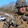 10th Marine Regiment HIMARS Firing Excercise A U.S. Marine officer in full combat gear is speaking or giving instructions in an outdoor field environment. He is wearing MARPAT woodland digital camouflage, a tactical vest with a holstered pistol, and a combat helmet with a mounted device. A second Marine is partially visible in the background, along with a white pickup truck and bare winter trees.