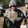 Three military personnel in U.S. Marine Corps MARPAT digital camouflage uniforms are gathered together, appearing to be in a briefing or discussion. The central figure wears a plate carrier vest with two visible stars indicating a Major General rank, tactical gloves, and a helmet-mounted device. The setting appears to be an outdoor training or operational environment with sandy terrain and trees in the background.