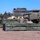 Military personnel in camouflage uniforms are handling large rocket pod containers near a multiple launch rocket system (MLRS) vehicle. The scene appears to be a loading or resupply operation at an outdoor training or operational area. The green ammunition pods bear standard military handling markings and are being positioned for loading onto the launcher.