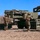 10th Marine Regiment HIMARS Firing Excercise Military personnel in combat uniforms and helmets are operating what appears to be M270 Multiple Launch Rocket System (MLRS) or similar rocket artillery vehicles in an open field. Soldiers are engaged in what appears to be a reloading or maintenance operation, handling rocket pod containers. The setting appears to be a training exercise based on the organized, non-combat environment.