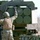 10th Marine Regiment HIMARS Firing Excercise A military service member in camouflage uniform and combat gear is working with a rocket artillery system mounted on a military vehicle. The soldier appears to be handling or inspecting a launcher pod containing multiple rocket tubes arranged in a 2x3 configuration. The setting appears to be an outdoor training or operational environment with sparse vegetation in the background.