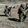 10th Marine Regiment HIMARS Firing Excercise Two military personnel in MARPAT camouflage uniforms and combat helmets are operating professional camera equipment with large telephoto lenses in an outdoor field setting. Both are kneeling on dry grass while photographing or filming a distant subject, with one camera mounted on a tripod equipped with a Rode microphone. The setting appears to be a military training area or exercise, indicated by boundary ropes, a tripod antenna structure, and tactical gear.