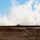 The image captures a rocket or missile launch from a ground-based system in an open, arid field. A bright launch flame is visible at the horizon line, with a dense white smoke plume trailing the projectile as it ascends steeply into a partly cloudy blue sky. The surrounding terrain is dry and flat with sparse low vegetation.