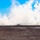10th Marine Regiment HIMARS Firing Excercise The image shows what appears to be a rocket or missile launch from a flat, open field with dry, sparse vegetation. A bright exhaust plume and smoke trail are visible rising steeply into a clear blue sky, accompanied by a large cloud of white smoke at ground level. The scene is consistent with a military weapons test or live-fire exercise.