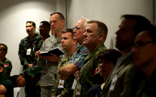 A group of military personnel from multiple nations are gathered in what appears to be a briefing room or conference setting. The individuals are wearing various military uniforms and civilian attire, with visible national insignia including a Canadian flag patch. They appear to be attentively observing something outside the camera frame, likely a presentation or briefing.