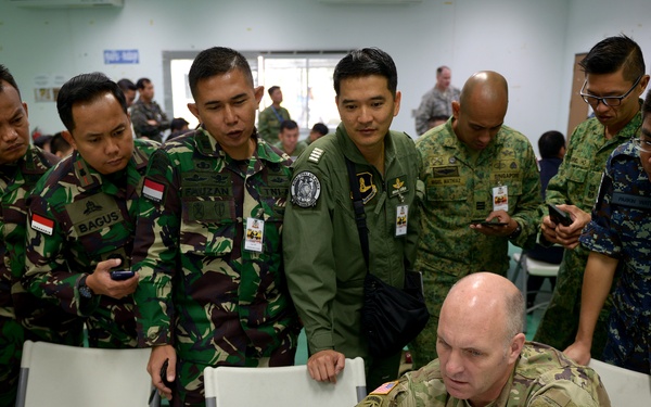 A group of multinational military personnel in various uniforms are gathered around a table observing a US Army soldier operating a laptop computer. The setting appears to be an indoor briefing or operations room, suggesting a joint military exercise or training event. Multiple national uniforms are visible, indicating an international coalition or multilateral military cooperation event.