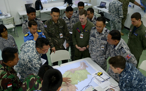 A multinational military planning session is taking place in what appears to be a briefing or operations room. Personnel from multiple nations, identifiable by their varied camouflage patterns and uniforms, are gathered around a table examining a large map alongside laptops and documents. The gathering suggests a joint military exercise or coordination meeting involving forces from several countries.