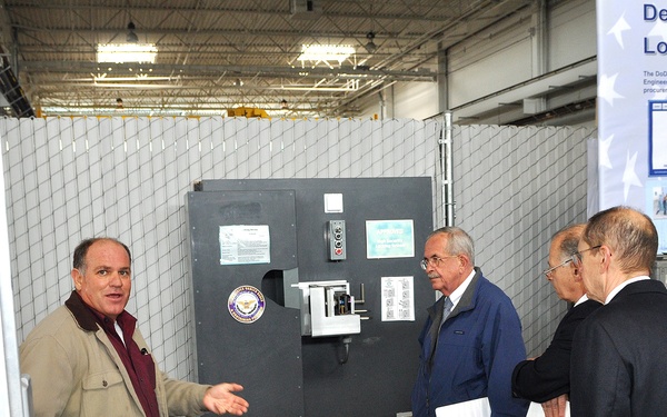 Three men are gathered in what appears to be a warehouse or facility demonstration area, where one man in a tan jacket is gesturing toward a large gray vault or security door mounted on a cart. A banner in the background references a 'Department of D... Lock Program,' suggesting this is a Department of Defense security equipment demonstration or briefing. Two men in formal attire, one holding white documents, appear to be receiving a tour or presentation.