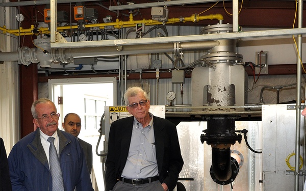 A group of men are gathered in an industrial or laboratory facility, listening to a presenter or guide who appears to be explaining equipment or processes. The setting includes visible industrial machinery, piping, and what appears to be a large concrete or stone sample on a table in the foreground. The occasion appears to be a formal tour or technical briefing.