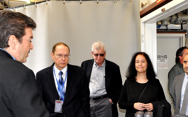 A group of approximately seven professionals are gathered in what appears to be an industrial or research facility, receiving a briefing or demonstration. A presenter on the left is gesturing toward metal components displayed on a table, which appear to be aerospace or engineering parts. The setting suggests a formal facility tour or technical review.