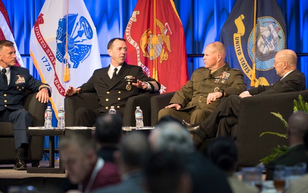 A formal military panel discussion is taking place on a stage before a large audience. Four individuals — three in military uniforms representing different branches and one in civilian attire — are seated in chairs engaged in conversation. The event appears to be an official military symposium or conference, indicated by the branch flags and organizational seal visible in the background.