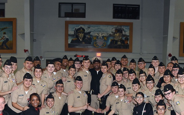 A large group of military cadets in tan/khaki uniforms poses for a formal group photograph with multiple trophies and awards displayed in front of them. The group includes both cadets and adult instructors/coaches wearing black jackets, suggesting this is a JROTC or similar military youth program. The setting appears to be a military facility or armory with large patriotic murals visible on the walls in the background.