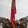 A U.S. Marine Corps change of command ceremony is taking place on a flight line or tarmac. Two senior Marines in MARPAT camouflage uniforms are exchanging a red unit guidon/colors, which is the central ritual of a change of command. Military aircraft, likely F-35s or similar jets, are visible in the background along with formation of Marines at attention.