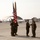 A military ceremony is taking place on an airfield tarmac, likely a change of command or award ceremony involving U.S. Marines in woodland MARPAT camouflage uniforms. A color guard holds the U.S. flag and what appears to be a unit guidon, while two personnel on the right appear to be exchanging or presenting an item. The background shows aircraft hangars with dome-style shelters, a control tower, and at least one military aircraft.