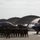 A group of military personnel in camouflage uniforms are lined up on an airfield tarmac between two stealth fighter jets, likely F-35s. Behind them are multiple large arched aircraft hangars. The scene appears to be a formal group photo or ceremonial gathering at a military air base.