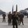 A U.S. Marine Corps officer addresses personnel during a formal ceremony on an airfield tarmac. The speaker holds a microphone and stands in front of a color guard bearing the U.S. flag and a unit guidon, with multiple Marines in MARPAT camouflage uniforms standing at attention. Two F-35 fighter jets are visible in the background, flanking the formation.