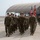 A formation of U.S. Marines in woodland MARPAT camouflage uniforms marches in disciplined ranks on what appears to be a military airfield tarmac. Color bearers carry the American flag and what appears to be a U.S. Marine Corps unit guidon. The scene suggests a formal military ceremony or parade, likely a change of command, activation, or similar official event.