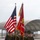 A U.S. Marine Corps color guard is conducting a formal parade or ceremony on what appears to be a military airfield tarmac. Four Marines march in formation carrying the U.S. national flag and a Marine Corps unit guidon, with additional Marines in formation behind them. The unit guidon partially reads 'Marine Attack Squ[adron]', indicating this is likely a USMC aviation unit ceremony.