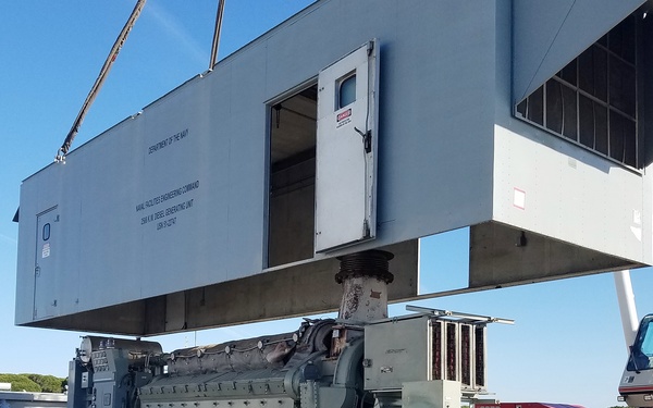 A large diesel generating unit belonging to the U.S. Department of the Navy / Naval Facilities Engineering Command is being lifted by a crane, with its protective enclosure housing being raised off the generator set below. The operation is taking place at what appears to be a port or naval facility on a clear, sunny day. A mobile crane operator is visible in the cab of a red and white crane vehicle on the right side of the image.