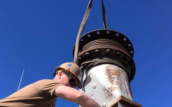 Two military personnel in uniform are working on a large industrial pump or mechanical equipment outdoors under a clear blue sky. One individual wearing a tan hard hat, safety glasses, and a brown t-shirt is actively manipulating a component on the machinery, while a second person in digital camouflage uniform assists. The equipment appears to be a large submersible or centrifugal pump showing significant rust and wear, being lifted or positioned using rigging straps.