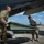 Two military personnel in camouflage uniforms are handling a large green metal ramp or loading panel beneath the wing/fuselage of a military transport aircraft on a flight line. The activity appears to be loading or unloading equipment from the aircraft. Additional military aircraft are visible in the background on the tarmac.