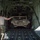 A military service member in multicam/OCP uniform is guiding a tan HMMWV (Humvee) being loaded into the cargo bay of a large military transport aircraft, likely a C-130 Hercules or C-17 Globemaster. The scene takes place on a flight line at what appears to be a military airfield. The loadmaster or crew member is directing the vehicle's positioning on the aircraft's cargo ramp.