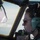 A female aviator is seated in a military aircraft cockpit, viewed in profile while in flight. She is wearing aviation headset equipment with a boom microphone, sunglasses, and what appears to be a flight suit. The cockpit window frames a sky and cloud backdrop, with a green-tinted heads-up display (HUD) visible to her left.