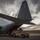 A United States Air Force C-130 Hercules transport aircraft is shown on a tarmac with its rear cargo ramp lowered. A tan/desert-colored HMMWV (Humvee) military vehicle is positioned at the base of the ramp, in the process of being loaded or offloaded. The scene is captured at dusk or dawn under dramatic overcast skies.