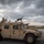 A tan/desert-colored military HMMWV (Humvee) is parked on what appears to be a tarmac or airfield surface. A soldier in combat gear is positioned in the rear of the vehicle, operating a radio with a tall whip antenna extended. The vehicle is equipped with a mounted crew-served weapon and bears a Roman numeral 'V' marking on its door.