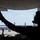 A silhouetted individual wearing a communications headset gives a thumbs-up gesture from inside or beneath a large aircraft, likely a military transport plane. The image is backlit, creating a dramatic silhouette effect against a bright airfield background. In the background, at least one military turboprop aircraft, consistent with a C-130 Hercules, is visible on the tarmac.