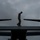A military service member in camouflage uniform walks along the top of what appears to be a CV-22 Osprey or similar tiltrotor/military aircraft. The figure is silhouetted against an overcast, dramatic sky, creating a striking contrast. The image appears to capture a pre-flight inspection or maintenance walk-around on the aircraft's fuselage.
