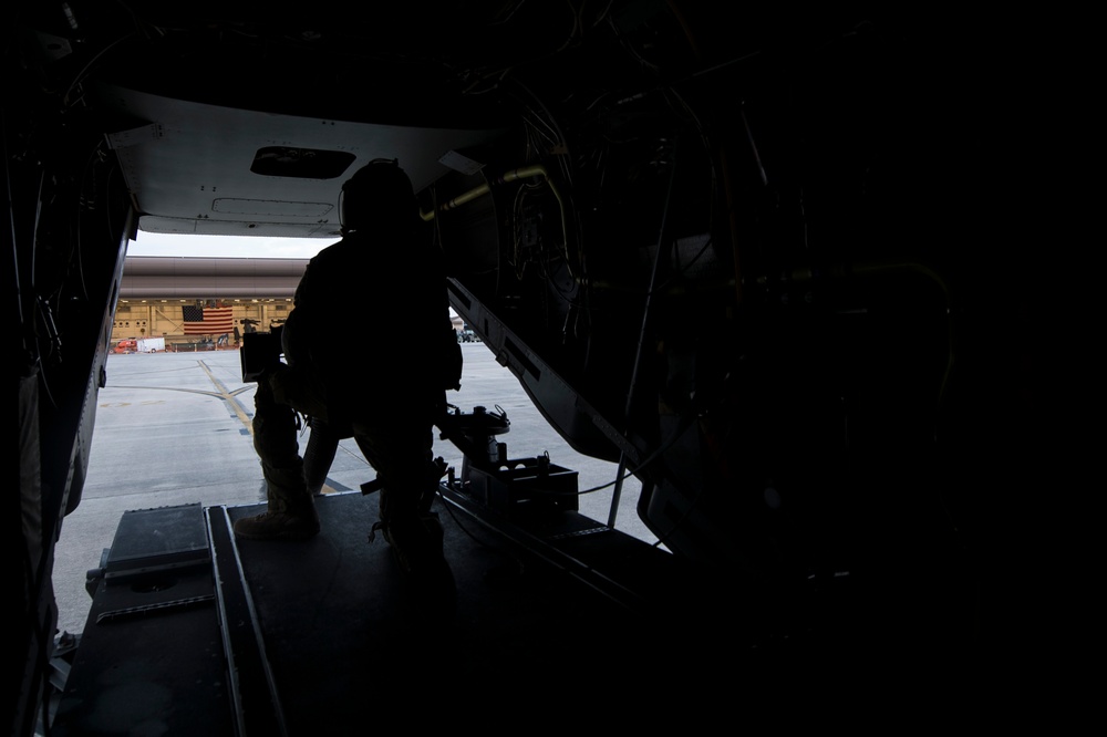 A silhouetted military personnel is positioned at the open rear ramp of a military aircraft, likely a cargo or transport helicopter/plane. An American flag is visible on a hangar building in the background, indicating a U.S. military base or airfield. The image is taken from inside the aircraft looking outward toward the tarmac.