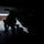 A silhouetted military personnel is positioned at the open rear ramp of a military aircraft, likely a cargo or transport helicopter/plane. An American flag is visible on a hangar building in the background, indicating a U.S. military base or airfield. The image is taken from inside the aircraft looking outward toward the tarmac.