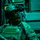 A military operator is seated inside what appears to be a military aircraft, illuminated by green ambient lighting. The soldier is equipped with full tactical gear including a helmet-mounted night vision device, body armor, and a communications headset. The background shows aircraft interior panels with visible safety markings.