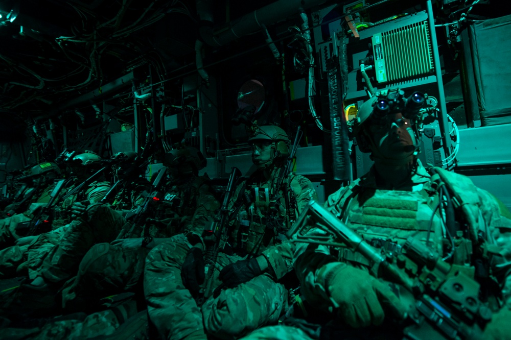 A group of heavily armed and equipped military personnel are seated inside what appears to be a military aircraft cargo hold, illuminated by green ambient lighting. The operators are wearing full tactical gear including helmets, body armor, night vision devices, and carrying assault rifles. The scene suggests a pre-mission insertion or infil aboard a transport or special operations aircraft.