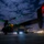 A tiltrotor aircraft, consistent with a V-22 Osprey, is photographed on a tarmac at night under a partially cloudy sky. The aircraft is illuminated by its own navigation lights (green and red) and ground-level landing lights, creating a dramatic low-light composition. A lit facility or fuel station is visible in the background to the right.