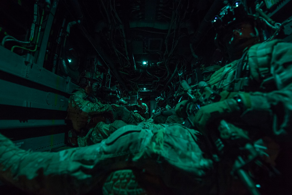 Military personnel in full combat gear are seated inside what appears to be a military transport aircraft or helicopter cabin, illuminated by dim green lighting. The soldiers are equipped with helmets, night vision devices, body armor, and tactical equipment. The image captures what appears to be an infil/exfil operation or transit during a nighttime mission.