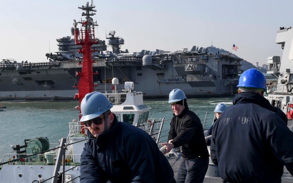 Naval personnel in blue hard hats and dark uniforms are handling mooring lines on the deck of a warship during a port operation. A large aircraft carrier is visible in the background, along with a tugboat alongside the vessel. The scene appears to take place in a naval harbor, likely during a docking or undocking evolution.