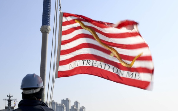 A naval personnel member wearing a white hard hat and dark jacket stands on the deck of a warship, facing a harbor with a coastal city skyline in the background. The First Navy Jack flag — featuring red and white stripes, a rattlesnake, and the motto 'Don't Tread On Me' — flies prominently from a mast. A tugboat is visible on the water in the middle distance.