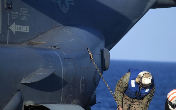 Two military personnel are securing a large military helicopter on what appears to be a naval vessel's flight deck at sea. One individual wearing Marine Corps camouflage uniform and a blue life vest is handling a chain attached to the aircraft, while a second person in dark clothing and a white helmet with red markings is working on a deck tie-down chain. Yellow wheel chocks are visible beneath the helicopter's landing gear.