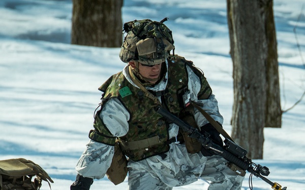 A soldier in winter camouflage gear is moving through a snow-covered wooded area while dragging a supply sled. The soldier is fully equipped with tactical vest, helmet with mounted accessories, and is carrying a rifle. The scene appears to be a cold-weather military training exercise or operation.