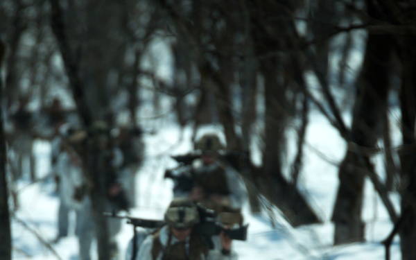 A column of military personnel in combat gear moves through a snow-covered forest in single file. The lead soldier is heavily laden with a large pack featuring winter camouflage covering, while soldiers behind carry rifles at the ready. The scene appears to depict a winter warfare training exercise or patrol operation.