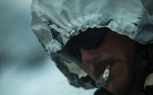 A military service member wearing winter camouflage gear and a helmet cover in a white/grey snow camouflage pattern is smoking a cigarette. Visible patches and insignia on the uniform suggest affiliation with the United States Marine Corps. The image is taken in what appears to be a cold, snowy environment with a shallow depth of field.