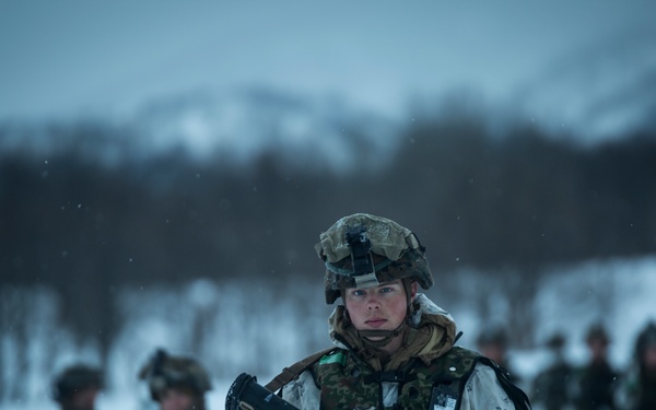 A group of military personnel conducting operations in a snowy, arctic or subarctic environment. The lead soldier in the foreground is equipped with full combat gear including a helmet with night-vision mount, body armor, and an assault rifle. Multiple soldiers are visible in the background, suggesting a unit-level patrol or training exercise in winter conditions.