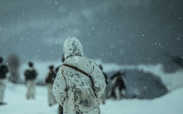 A military personnel in winter camouflage (white digital pattern) stands in the foreground during a snowstorm, facing away from the camera toward a group of similarly equipped soldiers in the background. The scene appears to be a cold-weather military training exercise or field operation conducted in heavy snowfall. Multiple personnel and what appears to be a low-profile shelter or tent structure are visible in the mid-ground.
