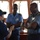 The image shows a group of military or naval personnel engaged in a discussion aboard what appears to be a ship's bridge or command area. A female officer in dark blue uniform and cap is speaking with two male officers wearing light blue uniforms with shoulder insignia and name badges. The setting suggests an official meeting or briefing between personnel from potentially different naval organizations.