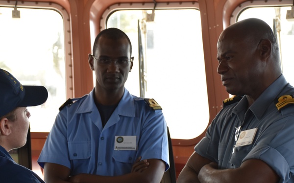 The image shows a group of military or naval personnel engaged in a discussion aboard what appears to be a ship's bridge or command area. A female officer in dark blue uniform and cap is speaking with two male officers wearing light blue uniforms with shoulder insignia and name badges. The setting suggests an official meeting or briefing between personnel from potentially different naval organizations.