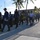 A group of military and law enforcement personnel in various uniforms are walking together across a paved parking area or roadway. The scene appears to be a joint multinational exercise or official visit, with personnel wearing different service uniforms including what appears to be U.S. Coast Guard and other national military uniforms. The setting is a tropical coastal location with palm trees, a multi-story building, and a communications tower visible in the background.
